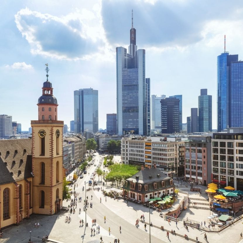 Luftaufnahme einer Stadt mit historischem Uhrturm neben modernen Glaswolkenkratzern, darunter ein Platz mit Cafés