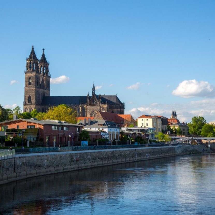 Blick auf eine gotische Kathedrale mit Doppeltürmen am Flussufer, umgeben von Stadtgebäuden entlang einer steinernen Uferpromenade unter klarem Himmel