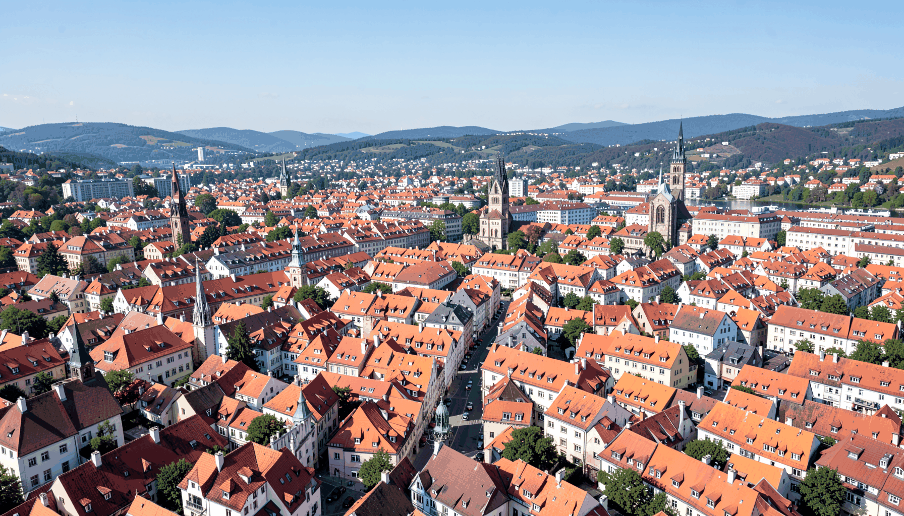 Weitwinklige Luftaufnahme einer dicht bebauten europäischen Stadt mit roten Ziegeldächern, Kirchtürmen und sanften Hügeln im Hintergrund unter klarem Himmel